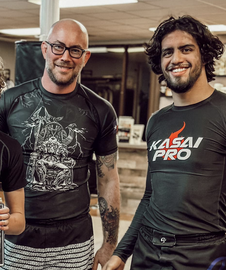 Two smiling male martial artists in compression shirts posing together inside a martial arts gym.